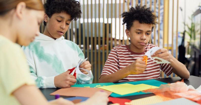 Idea Generation - Group of multiethnic children sitting at table and making artworks with colorful paper and scissors in light room