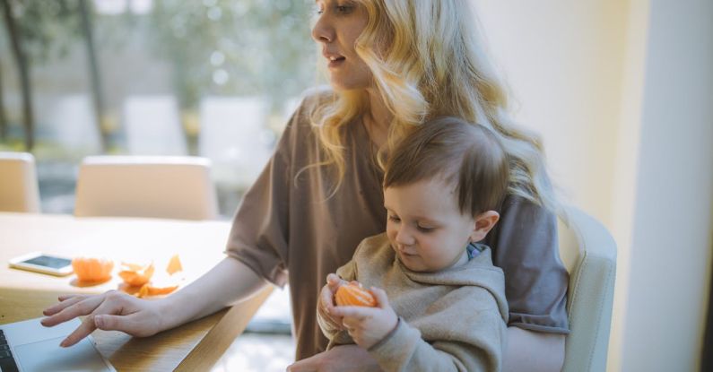 Managing Multitasking - Photo Of Woman Carrying Her Baby While Working On Her Laptop