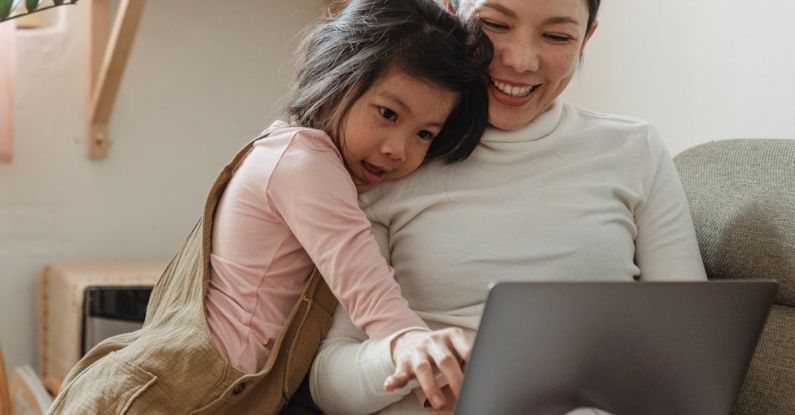 User-generated Content - Cheerful smiling Asian woman browsing modern netbook while hugging with cute content daughter on comfy sofa in cozy living room